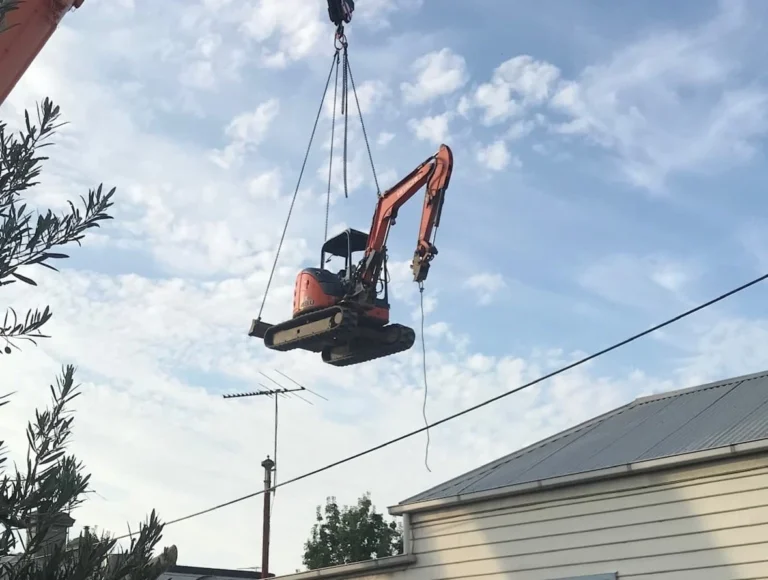 Excavator being lifted over a house by a crane for a tight access site cut.